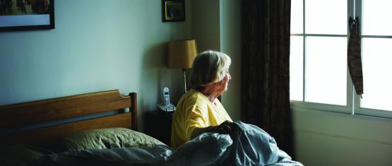 An older woman sitting on bed and looking outside the window
