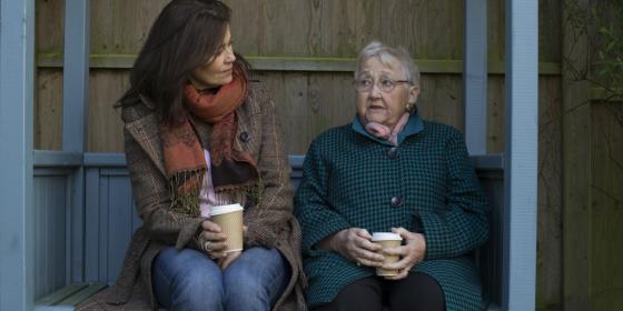 A woman and an older woman sit on a garden bench, talking
