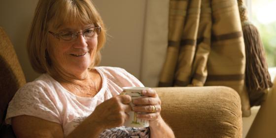 Woman in armchair holding a mug. She is wearing a white t-shirt and is facing towards the camera, looking off to the right. She is smiling.