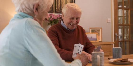 A man and a woman sitting together playing cards. The woman is wearing a blue cardigan and is sat with her back partially to the camera. The man is facing the camera and is laughing.